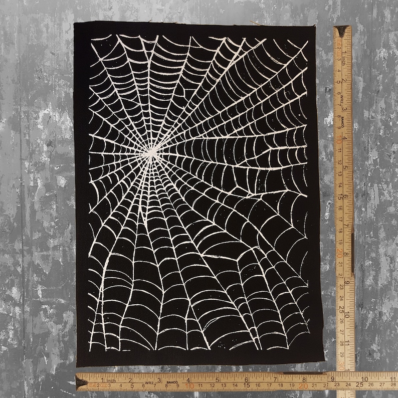 K&ouml;nnte beinhalten: Schwarzer Stoff-Patch mit einem wei&szlig;en Spinnennetz-Design. Das Netz ist detailliert und filigran und strahlt von der Mitte aus.