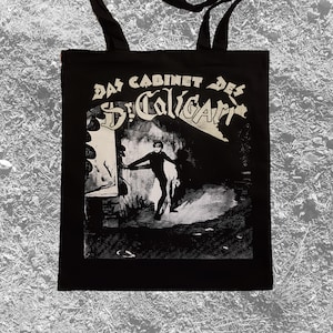 May include: Black tote bag with white text that reads "Das Cabinet Des Dr. Caligari". The bag features a black and white image of a person in a dark setting.