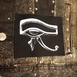 Puede incluir: Una chaqueta vaquera negra con un parche blanco del ojo de Horus cosido en la parte delantera. La chaqueta tiene tachuelas plateadas alrededor del parche y en la parte delantera y trasera de la chaqueta.