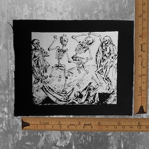 May include: Black and white embroidered patch featuring a scene of five skeletons dancing. The patch is sewn onto a black fabric background.