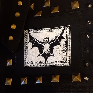 Op de afbeelding: Zwart denim jack met zilveren studs en een witte patch met een zwart-witte illustratie van een vleermuis.