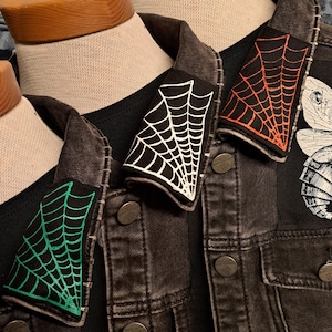 May include: Close-up of three black denim jackets with spiderweb patches in white, orange, and green. The jackets are displayed on mannequins, showcasing the patches' placement on the collar.