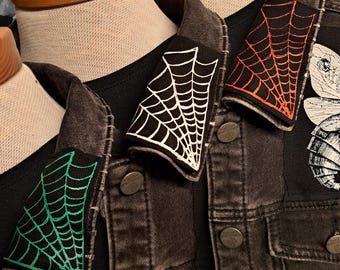 May include: Close-up of three black denim jackets with spiderweb patches in white, orange, and green. The jackets are displayed on mannequins, showcasing the patches' placement on the collar.