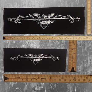 May include: Two black rectangular patches with a white graphic design. The design features a central grotesque face with bat wings and clawed hands on either side. The patches are approximately 6 inches long.