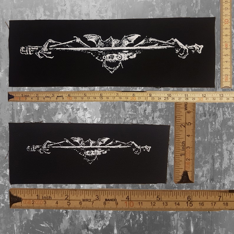 May include: Two black rectangular patches with a white graphic design. The design features a central grotesque face with bat wings and clawed hands on either side. The patches are approximately 6 inches long.