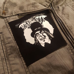 Op de afbeelding: Een zwart-wit patch met een grijnzende figuur met hoge hoed en uitgestrekte handen. De patch is bevestigd op een grijze spijkerjas met een rits en een zilveren stud op de schouder.