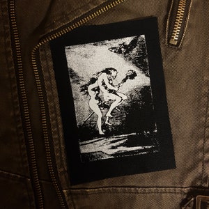 Puede incluir: Parche bordado en blanco y negro con una imagen estilizada de dos figuras, una de ellas sosteniendo un bastón, sobre un fondo nublado.