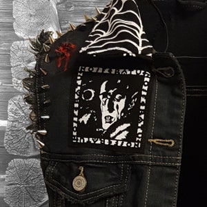 May include: A black denim jacket with a black and white spiderweb design on the collar. The jacket has silver studs on the shoulders and a black and white patch with a vampire image and the text "KULTUR REVOLUTION".