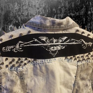 May include: A light-wash denim jacket with silver cone studs along the shoulders and collar. A large black patch on the back features a white graphic of a bat, skull, and crossed bones. The word "LIFE" is also visible.