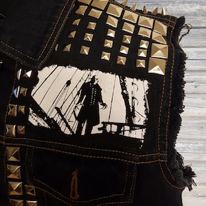 May include: A black denim vest with silver pyramid-shaped studs and a black and white patch depicting a figure in a long coat standing on a ship's deck.