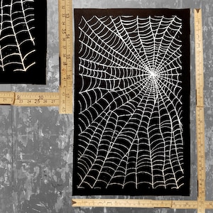 K&ouml;nnte beinhalten: Ein schwarzer Stoff-Patch mit einem wei&szlig;en Spinnennetz-Design. Das Netz ist detailliert und filigran und strahlt von der Mitte aus.