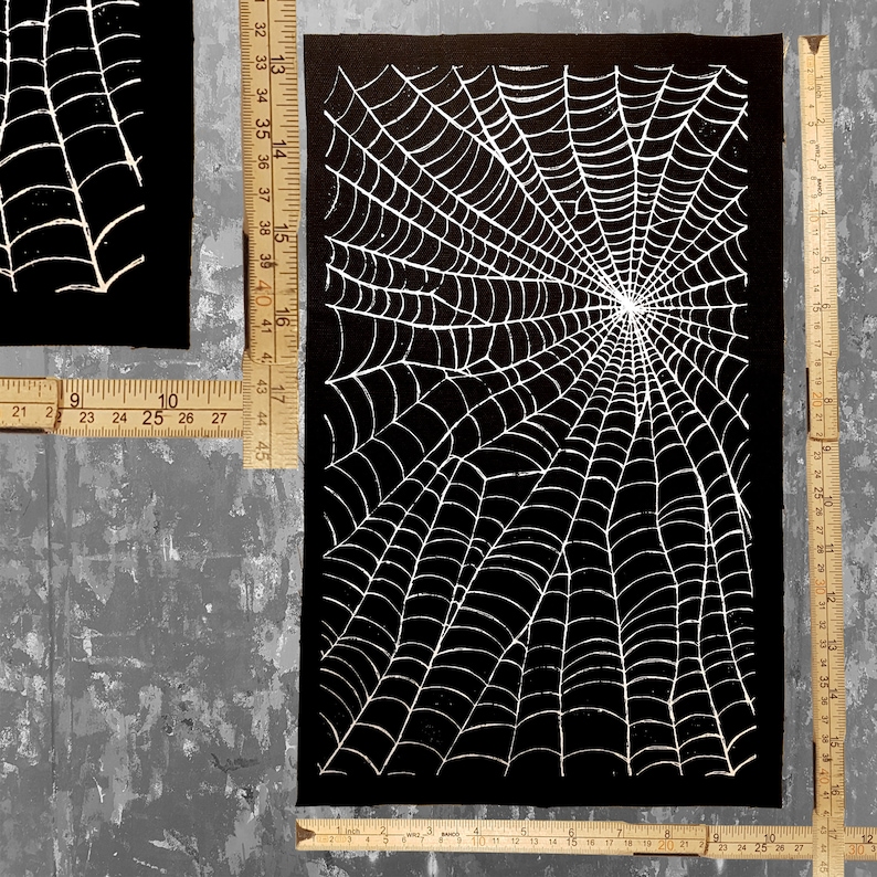 K&ouml;nnte beinhalten: Ein schwarzer Stoff-Patch mit einem wei&szlig;en Spinnennetz-Design. Das Netz ist detailliert und filigran und strahlt von der Mitte aus.