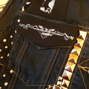 May include: Close-up of a black denim vest with gold stitching and metal studs. The vest features a small pocket with a white graphic patch and a larger patch at the top. The vest has a punk rock aesthetic.