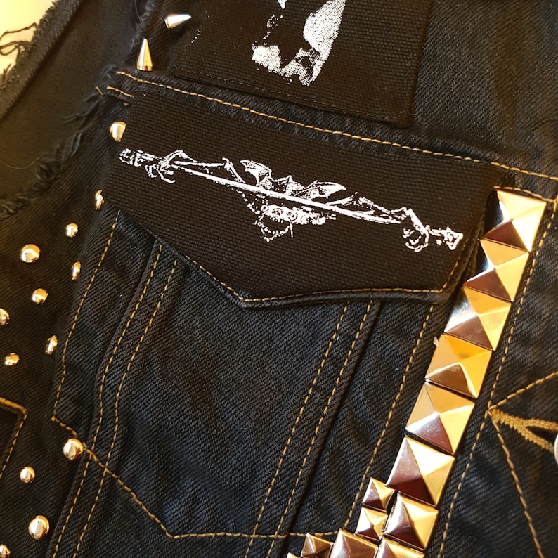 May include: Close-up of a black denim vest with gold stitching and metal studs. The vest features a small pocket with a white graphic patch and a larger patch at the top. The vest has a punk rock aesthetic.