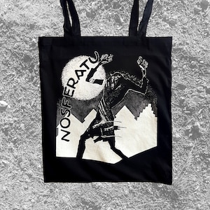 May include: Black tote bag with a white graphic of a vampire-like creature with the word "NOSFERATU" printed vertically on the left side of the image.