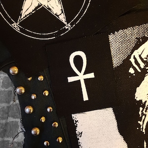 Pode incluir: Um remendo de tecido preto com um símbolo Ankh branco. O remendo é costurado em um fundo de tecido preto com tachas prateadas.