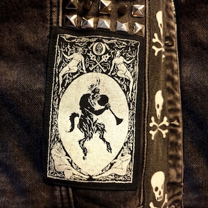 Peut inclure: Une veste en jean noir avec un écusson rectangulaire présentant une illustration blanche d'un satyre cornu jouant de la corne dans un cadre ovale, entouré de quatre figures. La veste comporte des clous pyramidaux argentés et un motif de crânes.