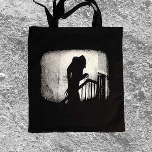 May include: Black tote bag with a white silhouette of a figure climbing a staircase. The figure is in a dark, shadowy setting.