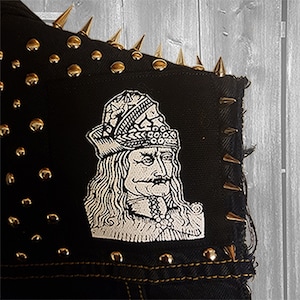 Può includere: Una giacca di jeans nera con borchie dorate e una toppa ricamata bianca di un uomo con una corona e un baffo.