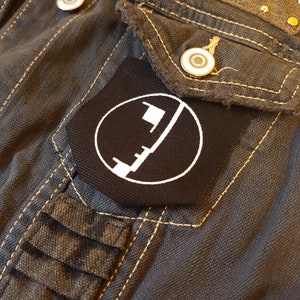 Peut inclure: Un écusson brodé noir et blanc avec un cercle et une ligne diagonale à l'intérieur. L'écusson est cousu sur une veste en jean gris foncé.