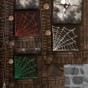 May include: Close-up of a dark denim jacket adorned with patches. The patches feature spiderweb designs in red, white, and green. The jacket has metal buttons and a faded, distressed appearance, suggesting a vintage or punk aesthetic.