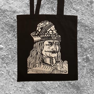 May include: Black tote bag featuring a detailed, stylized portrait of a historical figure with a mustache and ornate headwear. The image is in white against the black fabric. The bag has two black handles.