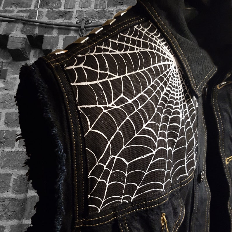 K&ouml;nnte beinhalten: Eine schwarze Jeansjacke mit einem wei&szlig;en Spinnennetz-Design auf der Schulter. Die Jacke hat silberne Nieten um den Kragen und die Schulter.