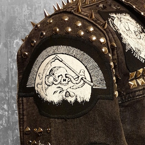 Op de afbeelding: Een donkergrijze spijkerjas versierd met zilverkleurige metalen spikes en studs. Een ronde zwart-witte patch toont een schedel met een slang door de oogkassen, een zeis en een kleine plant. Een andere patch is gedeeltelijk zichtbaar aan de bovenkant.