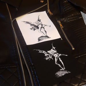 Könnte beinhalten: Eine schwarze Lederjacke mit zwei weißen Patches, die auf der Vorderseite angenäht sind. Die Patches zeigen eine Schwarzweißillustration einer geflügelten Kreatur, die ein Buch trägt.
