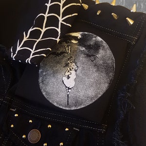 Könnte beinhalten: Schwarze Jeansjacke mit goldfarbenen Nieten und einem ausgefransten Saum. Sie hat einen runden Aufnäher mit einer Graustufen-Illustration einer Figur, die an einem Mond hängt. Ein schwarz-weißes Spinnennetz-Muster ist sichtbar.