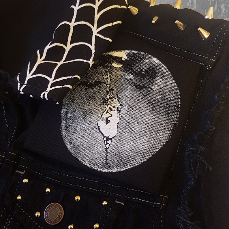 May include: Black denim jacket with gold-colored studs and a distressed hem. Features a round patch with a grayscale illustration of a figure hanging from a moon. A black and white spiderweb pattern is visible.