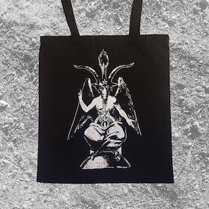 May include: Black tote bag with a white print of a horned figure with wings, sitting on a throne. The figure has a pentagram on its forehead and the words "Solve" and "Coagula" written on its arms. The text at the bottom reads "Cliphas Levi Del".