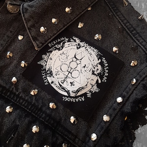 May include: Black denim jacket with silver studs and a black patch with a white design. The patch features a circle with a triangle inside, a cross, and the words "ASPARADEC", "AMASARAC", "AKIBECC", and "BERKAIAC".