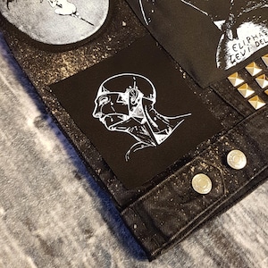 May include: A black denim jacket with a white anatomical drawing of a head on a patch. The jacket has a distressed look with white speckles throughout. The patch is sewn onto the jacket with silver studs around the edges.