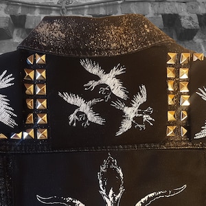 Puede incluir: Chaqueta vaquera negra con tachuelas plateadas y pájaros bordados en blanco. La chaqueta tiene un aspecto desgastado con salpicaduras de pintura blanca.