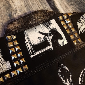 May include: A black denim jacket with various patches, including a black and white patch of a figure reaching out of a window. The jacket is decorated with silver pyramid-shaped studs.