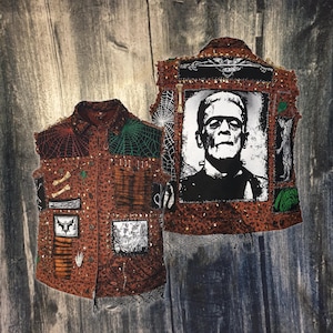 Puede incluir: Chaleco marrón con varios parches, incluyendo telarañas, huesos y un murciélago. El chaleco presenta un estampado de leopardo y una gran imagen en blanco y negro del monstruo de Frankenstein en la espalda. El chaleco está decorado con tachuelas y bordes deshilachados.