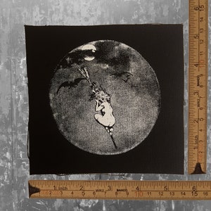 May include: Black and white patch featuring a witch riding a broom against a full moon backdrop. The witch is depicted in a seated position, with bats flying in the sky. The patch is square-shaped.