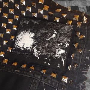 Puede incluir: Chaqueta vaquera negra con tachuelas piramidales plateadas y un parche negro y blanco con una imagen de una escena de una película o libro. El parche está cosido en la chaqueta.