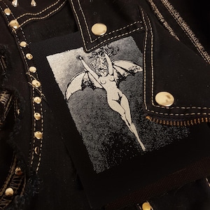 Puede incluir: Parche termoadhesivo en blanco y negro con una figura desnuda con alas, que se asemeja a un murciélago o un ángel, sobre un fondo oscuro.