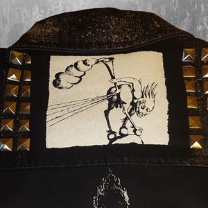 May include: Black denim jacket with a white patch featuring a black and white illustration of a creature with a horn and a cloud. The jacket has gold-coloured pyramid-shaped studs on the shoulders and sides.