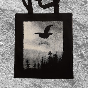May include: Black tote bag featuring a silhouetted bird in flight above a forest scene. The artwork is in a rectangular frame with a textured, cloudy background. The bag has black handles.