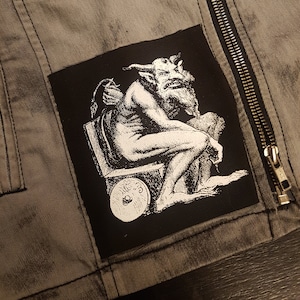 May include: Black and white patch featuring a detailed illustration of a horned demon seated on a throne. The demon is holding a staff and has a long beard. The patch is sewn onto a grey denim jacket.