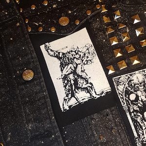 May include: A black denim jacket with silver studs and a white patch featuring a black and white illustration of a figure holding a weapon.