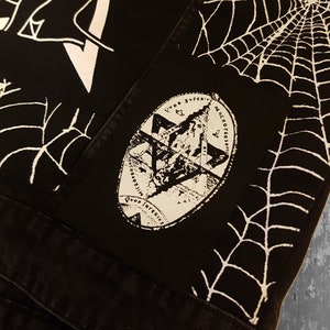 May include: Black denim jacket with a white spiderweb pattern and a white patch with a graphic design and the text "QUOD SUPERIUS SIC INFERIUS MICROCOSMOS MACROCOSMOS".