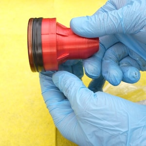 Op de afbeelding: Een rood en zwart cilindrisch object wordt vastgehouden door twee handen met blauwe handschoenen. Het object heeft een uitlopende basis en een zwarte band rond de bovenkant. De achtergrond is effen geel.