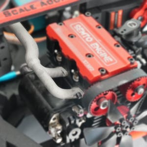 Op de afbeelding: Close-up van een modelmotor met een rode bovenkant met de tekst "SEMTO ENGINE". De motor heeft zwarte componenten, een grijze uitlaatpijp en een zwarte riem met rode tandwielen. Onderaan is een kleine ventilator zichtbaar.