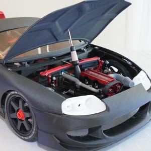 Könnte beinhalten: Ein detailliertes Modellauto mit geöffneter Motorhaube, das einen roten Motor und verschiedene mechanische Komponenten zeigt. Das Auto ist hauptsächlich schwarz mit weißen und roten Akzenten, die detaillierte Ausführung.