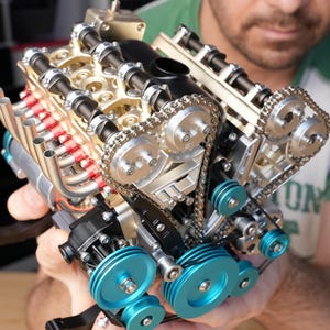 Könnte beinhalten: Ein detailliertes Motormodell mit goldenen, silbernen, schwarzen und blauen Komponenten. Es verfügt über Auspuffrohre, Zahnräder, Ketten und Riemenscheiben. Das Modell wird von einer Person gehalten.