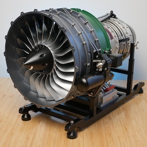 Puede incluir: Un modelo detallado de motor a reacción turbofan con un gran ventilador frontal de aspas plateadas y un cono central negro. La carcasa del motor es gris con detalles en verde, montada sobre un soporte de metal negro con el texto StirlingKit.